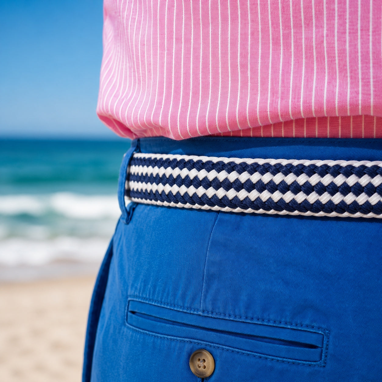 Person wearing a pink shirt and blue shorts with a striped belt, standing on a beach.